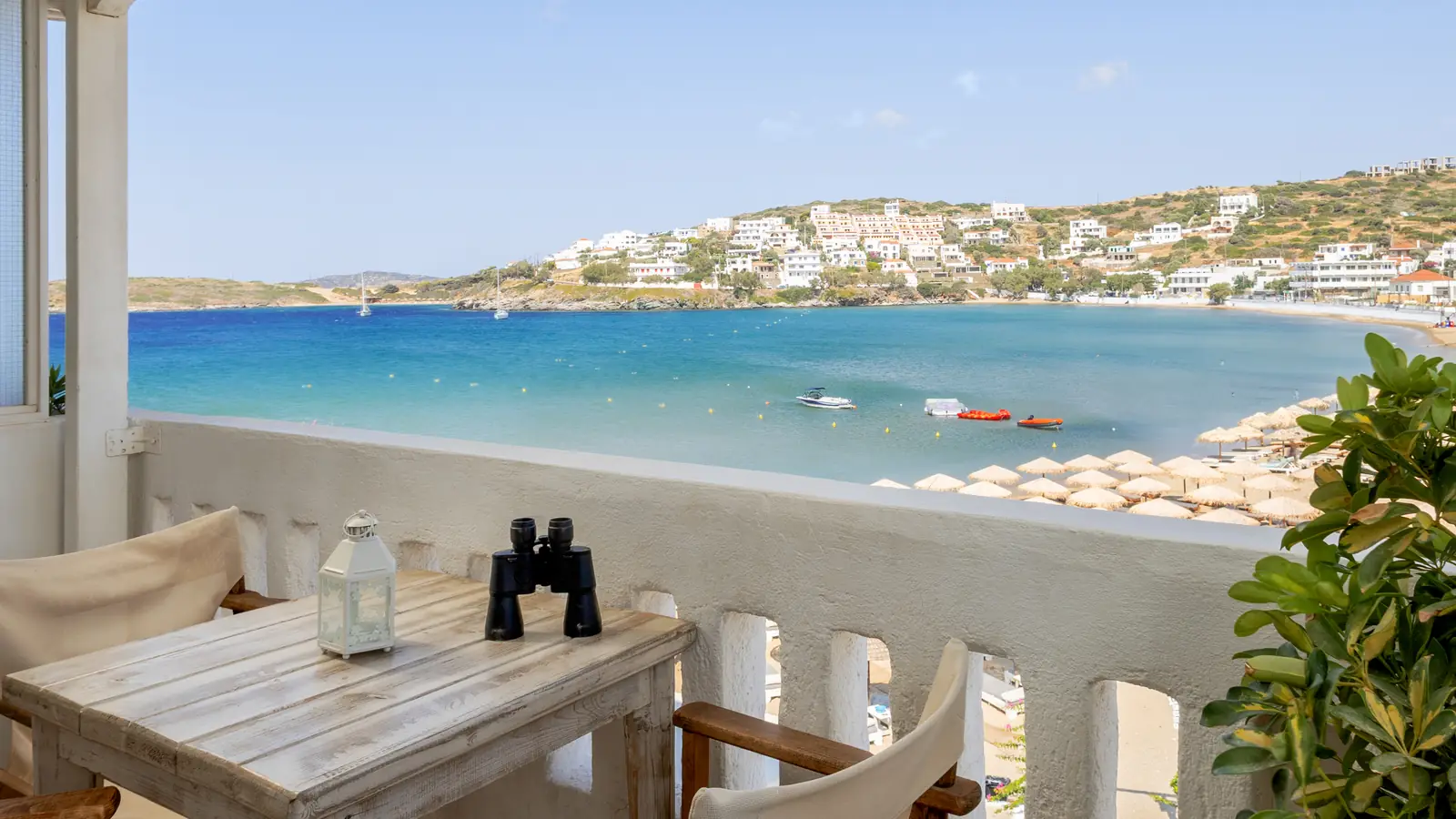 Τραπέζι μπαλκονιού με κιάλια, και πίσω ο όρμος του Μπατσίου με τις ομπρέλες, Άνδρος